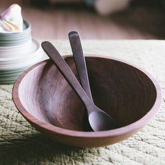 A black walnut wooden bowl with wooden salad tossers on top of a green cloth.