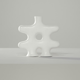 The Nedjma Vase by Schwung Design is a modern, abstract white ceramic piece with an irregular shape, round cutout center, and two short base supports. Handcrafted, it stands on a light surface against a plain gray background.
