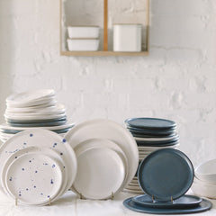 A variety of stoneware dinnerware including plates and bowls in different sizes, displayed on a table. Some plates have blue speckled patterns.