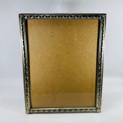 Vintage silver-tone metal 8x10 tabletop photo frame with ornate filigree leaf-and-scroll border, empty insert area shown, standing upright on a white background.