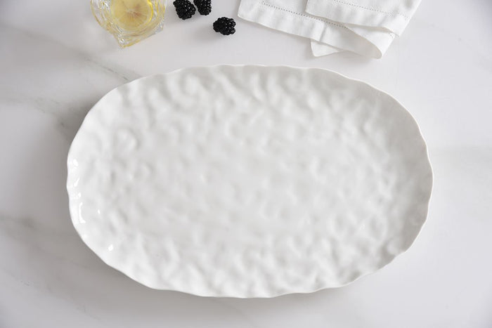 The PAMPA BAY Willow White Platter Oval Large, featuring a textured ceramic finish, rests on a white surface beside a folded napkin, a glass with lemon, and several blackberries.