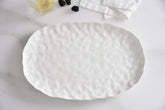 The PAMPA BAY Willow White Platter Oval Large, featuring a textured ceramic finish, rests on a white surface beside a folded napkin, a glass with lemon, and several blackberries.