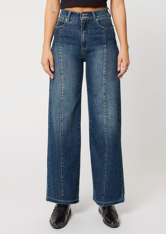 A woman wearing a pair of wide leg, high rise jeans in medium dark blue wash with seam down center leg.