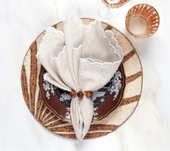 A table setting with a brown and white patterned plate, woven placemat, beige scalloped linen napkin held by the Kim Seybert Bois Napkin Ring Set of 4 - Brown & Gold, on marble beside two amber glasses.
