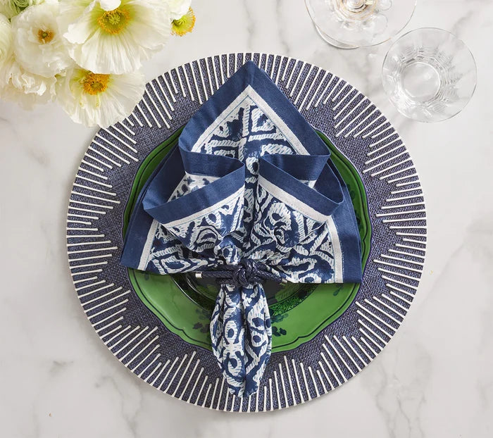 A blue and white napkin is folded on a green plate, set atop Kim Seybert’s Dream Weaver Placemat in navy & white. Nearby, two glasses and white flowers rest on the marble surface.