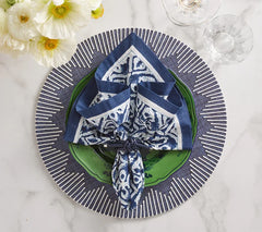 A blue and white napkin is folded on a green plate, set atop Kim Seybert’s Dream Weaver Placemat in navy & white. Nearby, two glasses and white flowers rest on the marble surface.