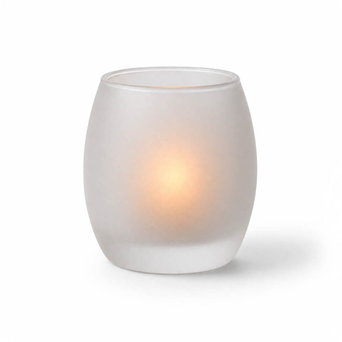 A glowing candle inside a frosted glass votive holder on a white background