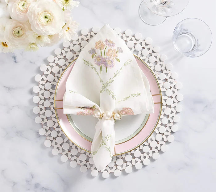 Elegant table setting with a white plate edged in pink and gold, topped with a floral embroidered napkin held by the Kim Seybert Pearl Drift Napkin Ring Set of 4 in Pearl & Gold. Clear glasses and white flowers rest on a marble surface.