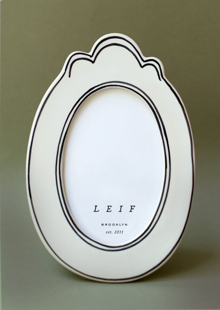 A white ceramic picture frame with ornate oval shape and thin handpainted border detail.