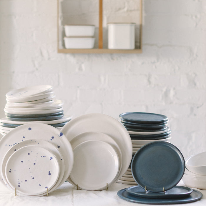 A variety of stoneware dinnerware including plates and bowls in different sizes, displayed on a table. Some plates have blue speckled patterns.