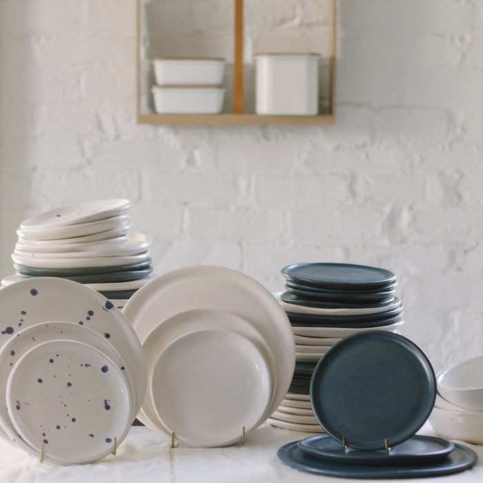 A variety of stoneware dinnerware including plates and bowls in different sizes, displayed on a table. Some plates have blue speckled patterns.